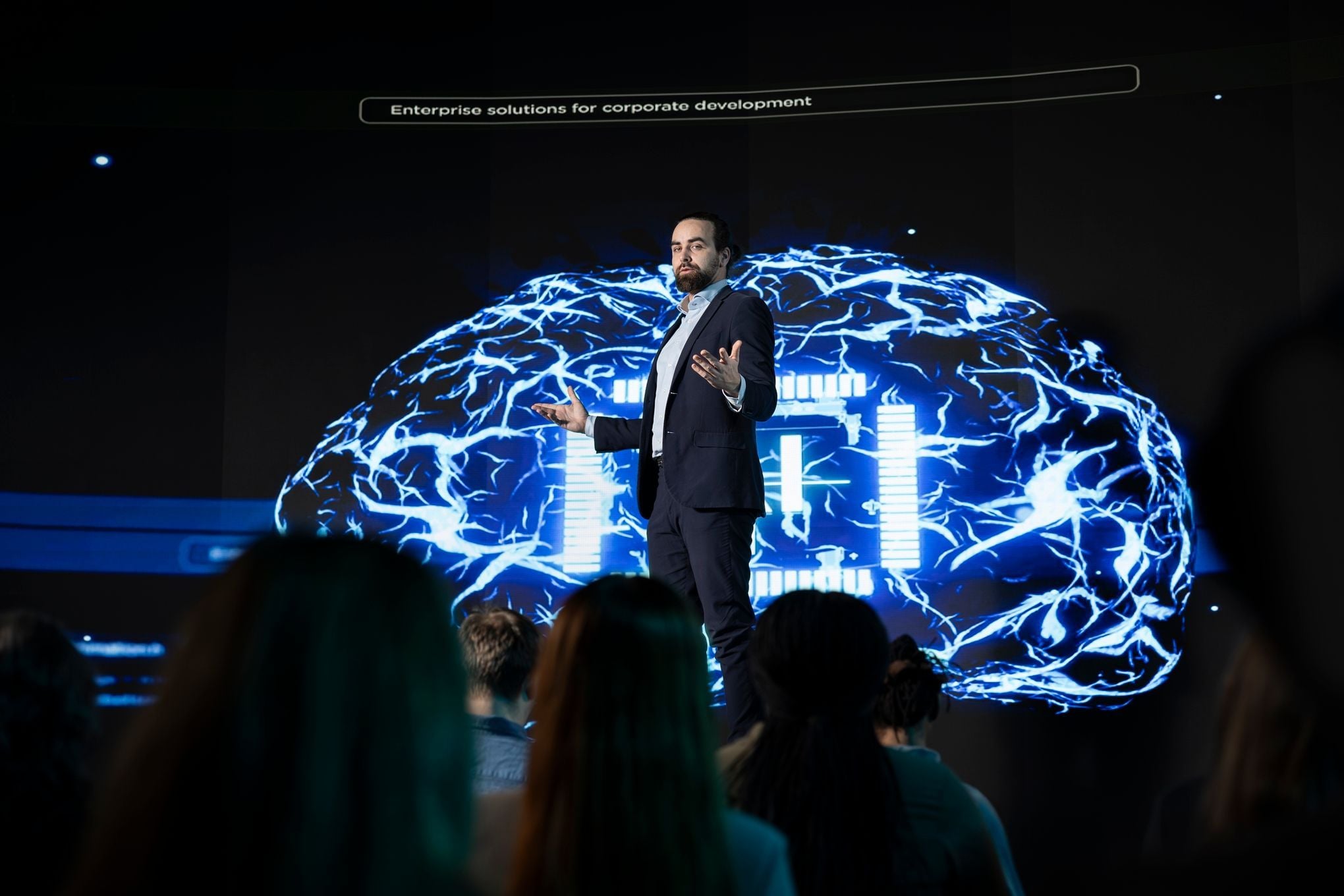 A professional speaker presenting "Enterprise solutions for corporate development" at an AI conference, standing in front of a large digital screen displaying a glowing blue neural network brain.