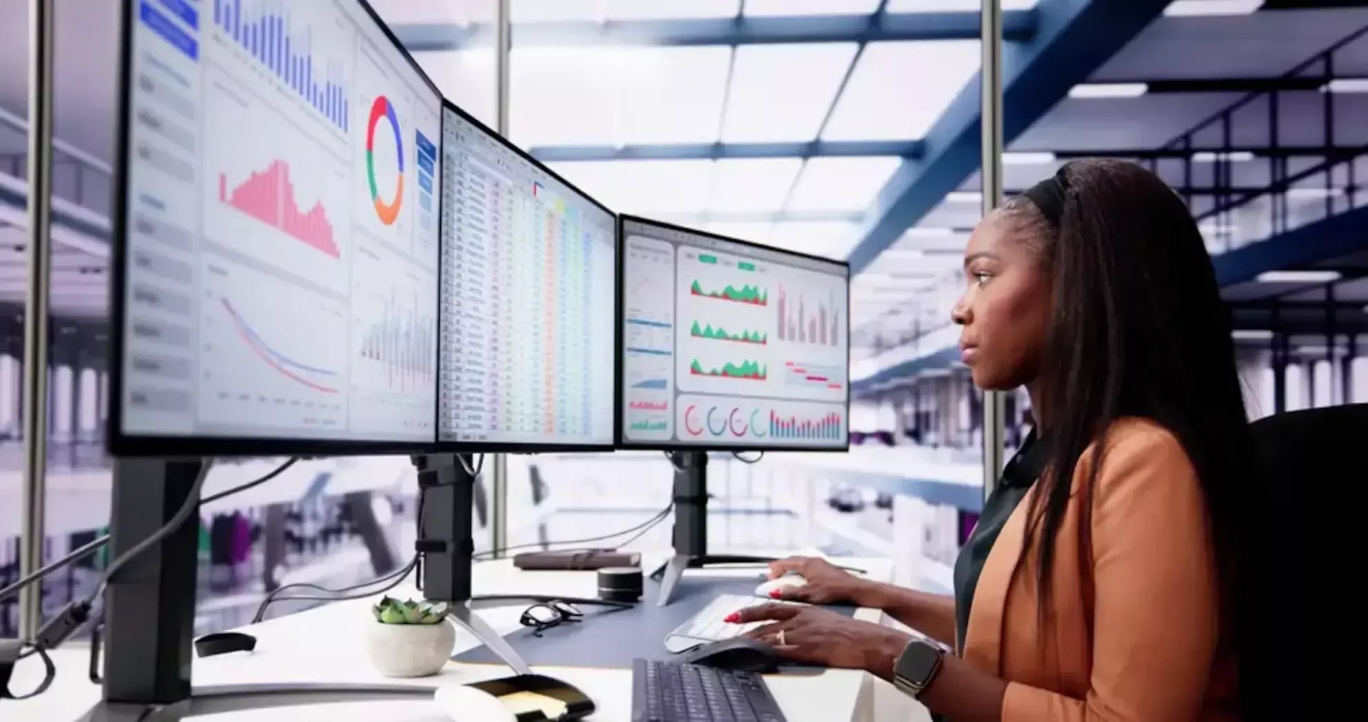 A professional female data analyst working at a modern workstation with a triple-monitor setup displaying complex data visualizations, charts, and spreadsheets in a bright office environment.