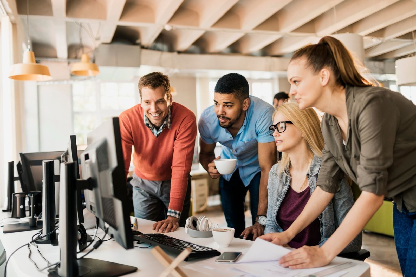 A diverse group of four professional developers and project managers collaborating around a computer monitor in a modern, brightly lit open-office environment.