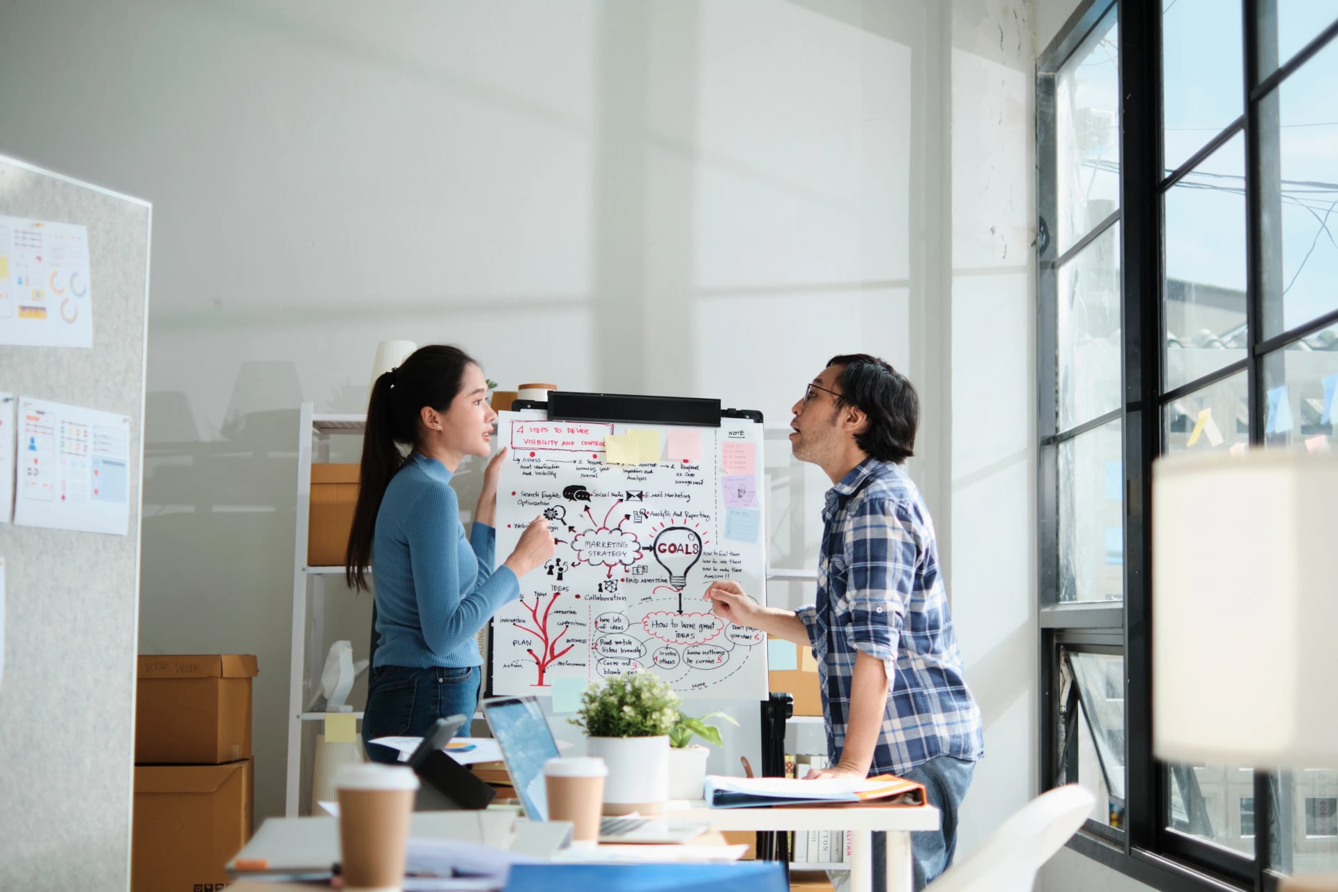 Two colleagues standing by a whiteboard easel in a sunlit office, discussing workflow strategies and project goals mapped out in diagrams.
