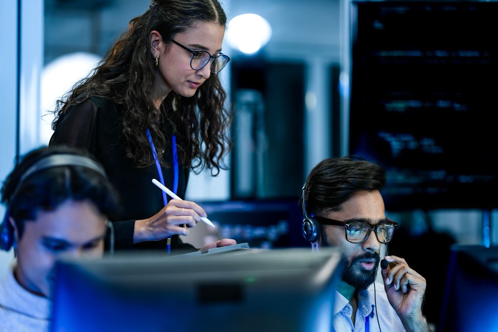 A tech lead with glasses using a stylus to collaborate with two developers at their workstations, reflecting the strategic restructuring toward smaller, highly experienced AI teams